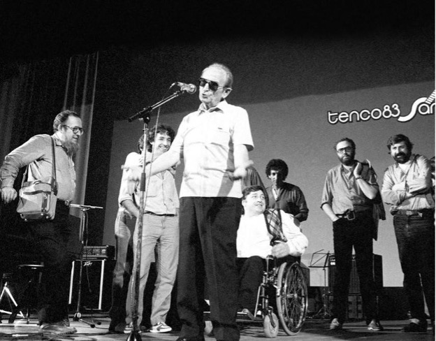 Chiusura dell'edizione 1983 del Premio Tenco. Al microfono Amilcare Rambaldi, storico “patron” della manifestazione; a sinistra Fausto Amodei, a destra Guccini, al centro Pierangelo Bertoli. "Mezze ali" (forse) David Riondino, - a sinistra. che ride - e Ivano Fossati, a destra con la giacca in spalla.
